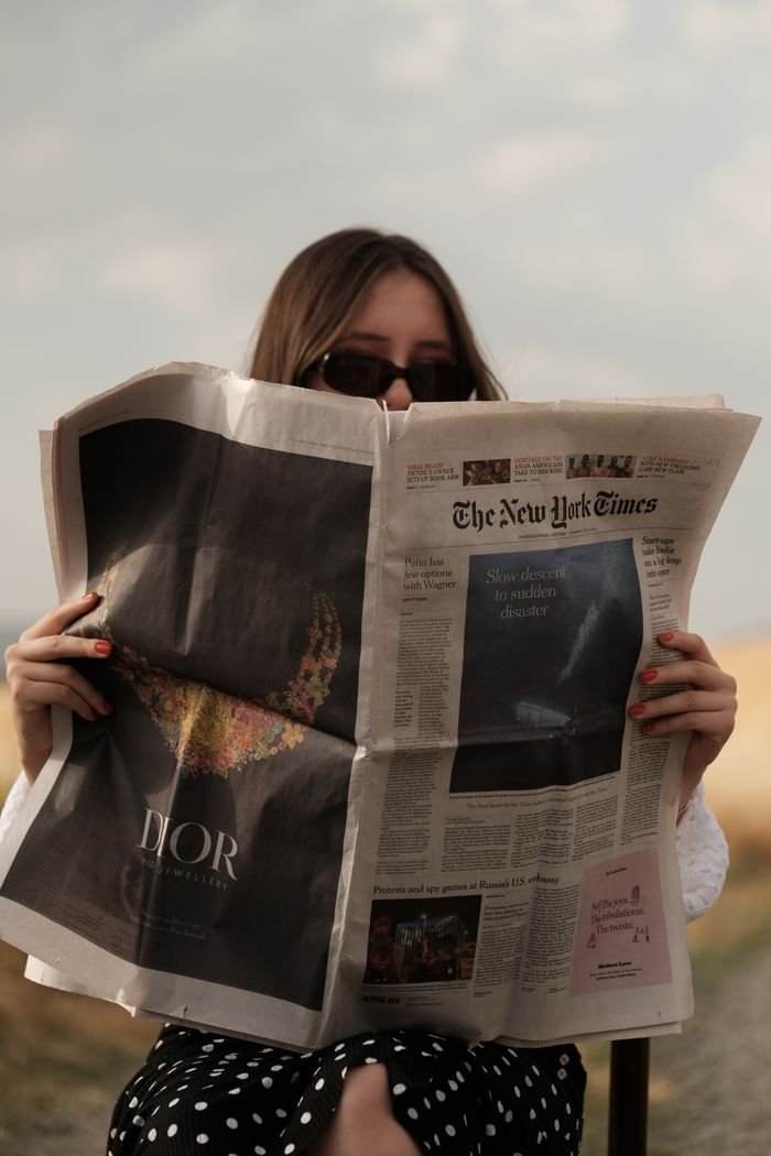 Services A woman in sunglasses reads The New York Times while seated outside, with a stylish fashion vibe.